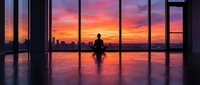 Silhouette of a person sitting cross-legged on a polished floor facing floor-to-ceiling windows overlooking a vibrant sunset cityscape with orange and pink hues reflecting on the glass and floor.