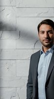 Middle-aged man with dark hair and beard wearing light blue shirt and charcoal blazer standing against white stone tile wall for corporate or professional business portrait use.