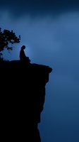 Silhouette of person sitting alone on cliff edge against dark blue night sky, with tree branch visible. Contemplative mood for concepts of solitude and meditation.