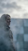 Middle-aged Caucasian woman in profile wearing gray coat against cloudy urban backdrop with blurred apartment buildings creating moody atmospheric portrait.