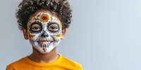 Young African American child wearing traditional Day of the Dead sugar skull face paint and orange t-shirt against light background, celebrating Mexican culture.
