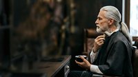 Senior man with long white hair and beard, wearing dark casual clothing, gesturing while speaking in an indoor setting. Suitable for business presentations, educational content, and communication concepts.