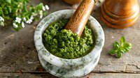 Fresh homemade basil pesto sauce in marble mortar with wooden pestle on rustic wooden table, garnished with herbs.