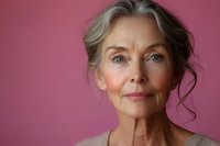 Elegant mature Caucasian woman with gray hair and natural makeup poses confidently against pink background, showcasing graceful aging and authentic beauty.