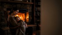 Young mother holding sleeping newborn baby by warm fireplace in cozy living room during evening hours creating peaceful family bonding moment at home.