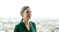 Mature woman with gray hair wearing green shirt gazing at cityscape view from high vantage point, contemplative expression, urban background for business and lifestyle concepts.