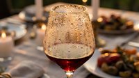 Chilled red wine glass with condensation drops on elegant dinner table with candles and food in soft bokeh background.