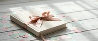 Elegant gift box with satin ribbon bow surrounded by scattered rose petals on white wooden surface for special occasions and celebrations.