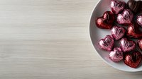 Heart shaped chocolate candies wrapped in red foil arranged in white ceramic bowl on light wooden surface, romantic holiday concept with copy space.
