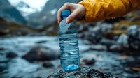 Person in yellow raincoat filling water bottle from mountain stream. Fresh water collection against blurred rocky landscape and flowing river background.
