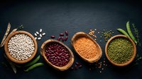 Assorted legumes and beans in wooden bowls on dark slate background with fresh herbs and spices for healthy cooking and nutrition concepts.