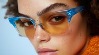 Young woman with freckles wearing stylish blue and yellow gradient cat eye glasses against light background, close up portrait with natural curly hair and orange sweater.