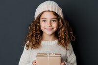 Young girl with curly brown hair wearing white knit beanie and sweater holds wrapped gift box against dark background, expressing genuine joy and anticipation.