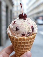 Cherry ice cream scoop in waffle cone topped with fresh maraschino cherry, creamy pink dessert with dark fruit pieces against blurred urban background.