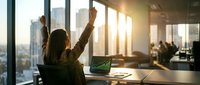 Successful businesswoman celebrating achievement in modern office with arms raised, city skyline view through windows during golden hour sunset.