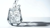 Clear water pouring into transparent glass with ice cubes creating splash and bubbles against white background for refreshment and hydration concepts.