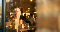 Young Asian woman smiling warmly while looking through cafe window at evening, golden hour lighting creating cozy atmosphere for hospitality and lifestyle concepts.