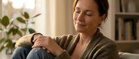 Young woman relaxing at home in cozy sweater, sitting peacefully on sofa with eyes closed in warm sunlight. Comfortable indoor lifestyle moment.