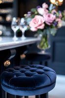 Elegant navy blue velvet tufted bar stool with pink roses and wine glasses in soft focus background creates luxurious restaurant atmosphere.