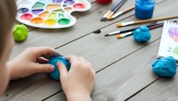 Child hands molding blue clay at wooden table with watercolor paints and brushes for creative art projects and educational activities.