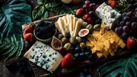 Gourmet cheese board with assorted cheeses, fresh berries, figs, olives, and grapes arranged on rustic wooden platter with tropical foliage.