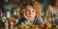 Young blonde curly-haired child resting chin on hand at kitchen table with warm lighting, contemplative expression for family lifestyle content.