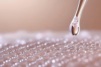 Transparent liquid droplet falling from dropper onto textured surface with water beads creating abstract pattern in soft pink tones. Macro photography captures surface tension.