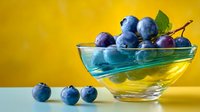 Fresh blueberries in glass bowl on vibrant yellow background, with scattered berries on table surface. Perfect for food blogs and healthy eating content.