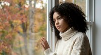 Young African American woman in cozy cream sweater looking thoughtfully out window during autumn season with colorful fall foliage background.