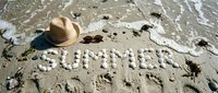 Word SUMMER written with seashells on sandy beach beside straw hat and sunglasses, with ocean waves in background.