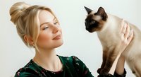 Young woman with blonde hair styled in a bun, fair complexion, wearing a dark green velvet top, holding and gazing affectionately at a seal-point Siamese cat against a white background. Suitable for pet care, animal companionship, and lifestyle content.