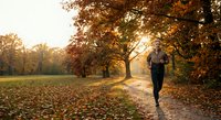 Young woman jogging through autumn park with golden sunlight filtering through colorful fall foliage on tree-lined path covered with fallen leaves.