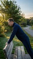 Middle-aged Caucasian man in sportswear doing push-ups on a park bench at sunset, with a winding path and green trees in background.