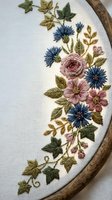 Delicate hand embroidered floral design with roses and cornflowers on white fabric in wooden hoop showcasing traditional needlework and textile art craftsmanship.