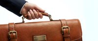 Businessman in dark suit holding brown leather briefcase handle against white background, representing business travel and professional work.