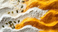 Vibrant orange pumpkin puree swirls on white flour background with scattered raw pumpkin seeds creating dynamic culinary composition for seasonal cooking.