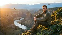 Man sitting on rocky cliff overlooking river canyon at sunset, contemplating nature landscape with mountains and flying birds in background.