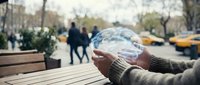 Person holding transparent glass sphere reflecting urban street scene with blurred pedestrians and yellow taxis in background on wooden table outdoors.