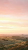 Rolling hills landscape at sunrise with soft pink and golden sky, gentle slopes covered in dry grass and vegetation, distant mountains on horizon, peaceful countryside scenery for nature and travel projects.