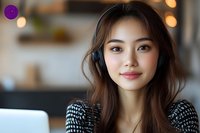 Young Asian woman wearing headphones smiles while working remotely from home office, dressed in casual sweater against blurred background with warm bokeh lights.