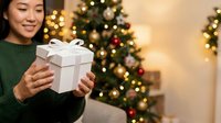 Young Asian woman holding white gift box with ribbon in front of decorated evergreen trees with warm golden lights and ornaments, celebrating holiday season and festive gift giving.