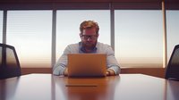 Professional man in glasses working alone at laptop in modern office conference room during sunset, warm lighting creates atmospheric business scene.