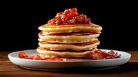 Stack of fluffy golden pancakes topped with red caviar on white plate against dark background, luxury breakfast fusion cuisine served on wooden table.