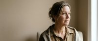Middle-aged Caucasian woman in beige shirt looking thoughtfully out window in warm natural light, contemplating life decisions and future plans.