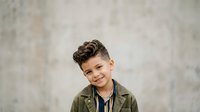 Young boy with dark hair and light skin tone wearing olive green jacket over navy shirt, smiling at camera against neutral gray textured background. Suitable for family, lifestyle, education, and children's content projects.