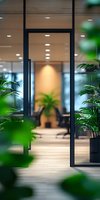 Modern office entrance with glass doors framed by lush green plants, featuring warm lighting and minimalist design. View through doorway shows contemporary workspace.