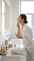 Young woman applying lip balm in modern bathroom mirror wearing white bathrobe during morning skincare routine with natural lighting and minimalist decor.