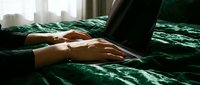 Young woman's hands typing on laptop while lying on luxurious dark green velvet bed, remote work and cozy home office concept.