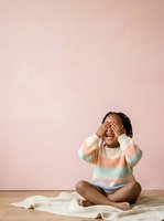 Young child, approximately 2-3 years old, of mixed ethnicity, sitting on wooden floor against soft pink wall wearing striped pastel sweater, covering face with hands in playful or shy gesture with crumpled newspaper beside them.