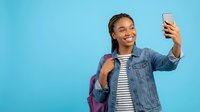 Happy young African American woman student taking selfie with smartphone wearing denim jacket and backpack on blue background for social media content.